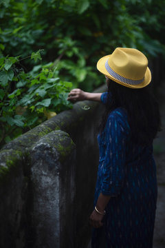 Backside view of an Indian woman standing on garden fencing wearing a blue dress and yellow western hat in a cloudy afternoon. Indian lifestyle