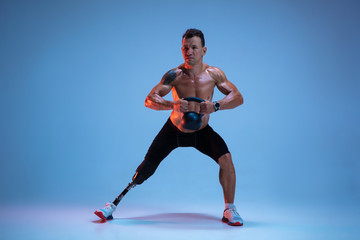 Fototapeta na wymiar Athlete with disabilities or amputee isolated on blue studio background. Professional male sportsman with leg prosthesis training with weights in neon. Disabled sport and overcoming, wellness concept.