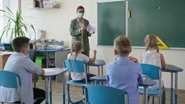 life after quarantine, teacher shows pupils cards with letters and numbers on lesson in classroom, masked children raises hand knowing the right answer