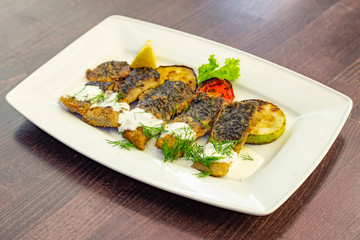 
Pieces of fried fish fillet with grilled vegetables (zucchini, tomato) decorated with lemon, dill and lettuce sprinkled with sour cream sauce. Fish on a white rectangular plate