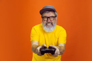 Tensioned gamer. Portrait of senior hipster man using devices, gadgets isolated on orange studio...