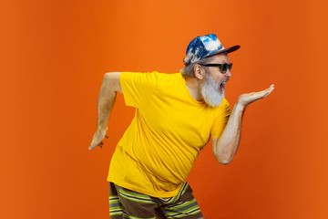 Crazy dance. Portrait of senior hipster man using devices, gadgets isolated on orange studio...