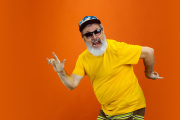 Crazy dance. Portrait of senior hipster man using devices, gadgets isolated on orange studio...
