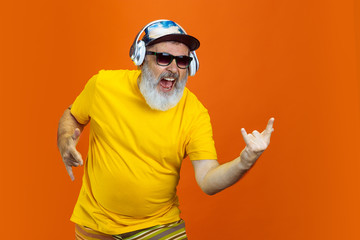 Crazy dance. Portrait of senior hipster man using devices, gadgets isolated on orange studio...