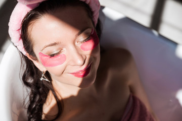 Fototapeta na wymiar Beautiful young woman in a pink cosmetic bandage and with pink birds under her eyes poses in front of the camera in a white ceramic bath in a sunny bathroom. Girl lies with closed eyes.