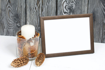Wooden photo frame with a white field for an inscription or a picture. Nearby is a candle in a candlestick with fir cones and dried rowan berries.