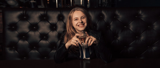 Wide angle shot of young girl sitting alone in a pub with a glass of whiskey in hand and smiling. Selective focus of woman in leather jacket looking at the camera. Image with copy space.