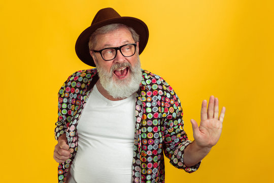 Stunning posing, greeting. Portrait of senior hipster man in eyewear isolated on yellow studio background. Tech and joyful elderly lifestyle concept. Trendy colors, forever youth. Copyspace for your