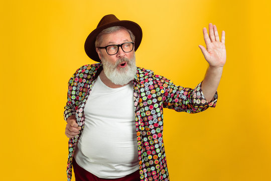 Stunning posing, greeting. Portrait of senior hipster man in eyewear isolated on yellow studio background. Tech and joyful elderly lifestyle concept. Trendy colors, forever youth. Copyspace for your