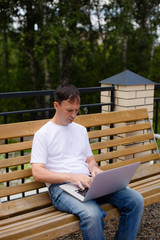 Fototapeta premium Portrait of a middle-aged man with laptop sitting on the bench. Man in white t-shirt and blue jeans with laptop sitting on the bench in the park.