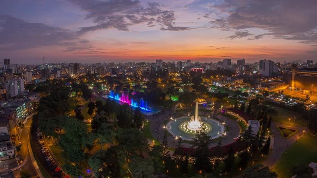 Aerial view to Park of the Reserve with magic water circuit biggest fountain complex day to night transition timelapse. Green lawn and trees. City skyline on a background
