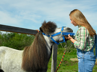 The young Caucasian woman and her gray pony are standing face to face near the hitching post.