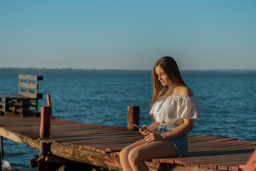 young blonde woman sitting on a pier using her cell phone