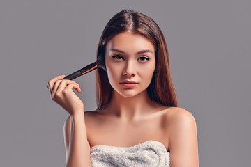 Cute attractive girl with a towel uses a brush for powder or blush isolated on a gray background. Skin care concept. Spa treatments, cosmetology, make-up.