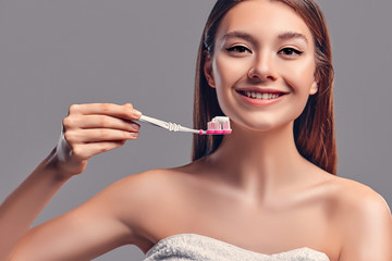 Young attractive brunette girl with loose hair uses a toothbrush and paste isolated on a gray background.