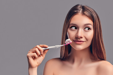 Young attractive brunette girl with loose hair uses a toothbrush and paste isolated on a gray background.