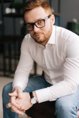 Serious young business man wearing stylish glasses in trendy clothing looking at camera while sitting indoors on chair in modern office. Portrait of bearded handsome gentleman wearing white shirt.