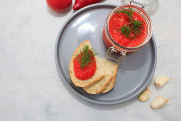 Ajvar (pepper mousse) in a jar and on gray plate with bread and gray backgground. Ajvar - delicious dish of red peppers, tomato and garlic.