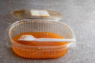 Open transparent plastic container with leftover food and a plastic spoon against a gray background, copy space