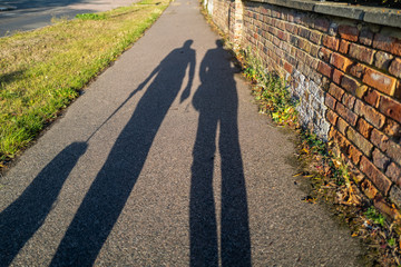 Long evening shadows of a man and woman walking the dog on a leash at golden hour dusk. They are walking on a pavement next to a grass verge and an interesting textures red brick wall.