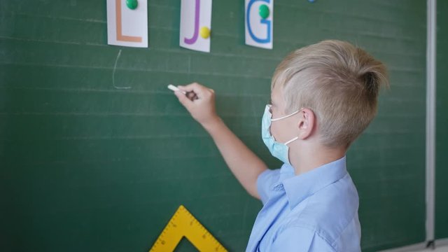schoolboy in a medical mask near blackboard writes in chalk the letters in classroom, school after quarantine and lockdown