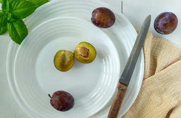 Several ripe plums and a knife with a wooden handle lie on a white porcelain plate on a light painted wood surface. Horizontal orientation, top view, selective focus.