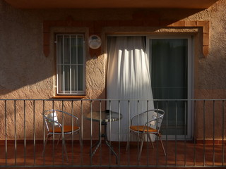 Terrace of a holiday appartment with a small round table and two chairs, open doors with white curtain blowing, Spain