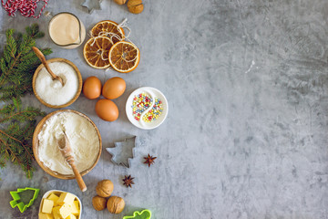 Culinary Christmas food background. Baking ingredients, flour, eggs, cookie cutters, fir branches, dried oranges, nuts, paper muffin molds on a dark table. Top view, copy space, flat lay