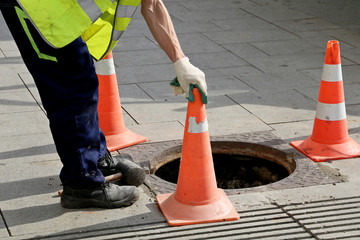 Worker over the open sewer hatch on a street near the traffic cones. Concept of repair of sewage,...