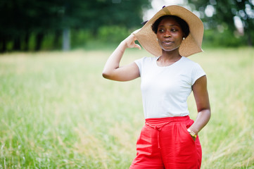 Portrait of gorgeous african american woman 20s wear in summer hat, red pants and white t-shirt, posing at green grass in park.