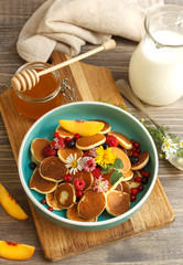 Mini pancakes decorated with fresh flowers and frozen berries in a blue bowl. On a wooden table with a jug of milk and honey in a jar
