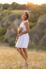 Fototapeta na wymiar Beautiful tender girl in a white sundress walks at sunset in a field with a spikelet bouquet.