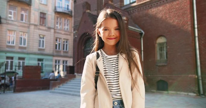 Portrait of joyful pretty cute Caucasian little girl smiling to camera outdoor. Nice beautiful schoolgirl in good mood standing in front of school alone with smile on face. Positive emotions concept