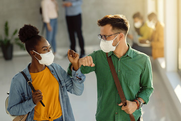 Happy college friends with face masks elbow bumping in a hallway.