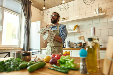 Young man, Italian cook wiping his hands with kitchen towel after cutting vegetables while preparing healthy meal, soup in the kitchen. Italian cuisine concept