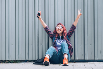 Beautiful modern smiling young female teenager in a checkered shirt and jeans with headphones and smartphone sitting near a wall on the street and Cheerful hands rising UP.Carefree student age concept