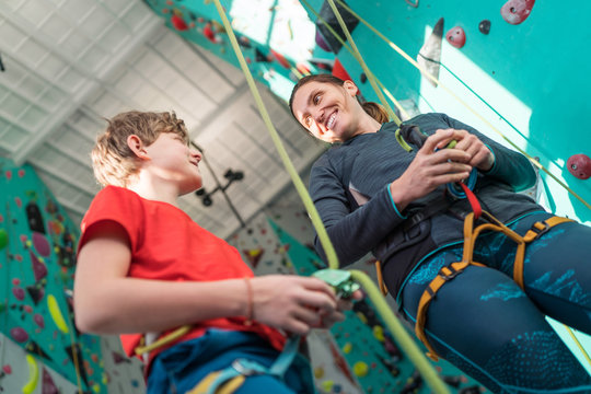 Mother and teenage son at indoor climbing gym near climbing wall. Boy preparing belaying his smiling mom. Happy parenting concept image.