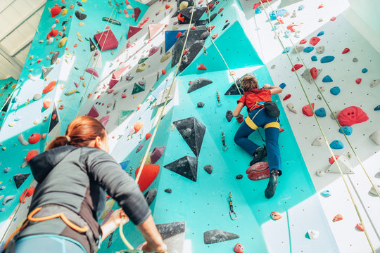 Mother and teenage son at indoor climbing wall gym. Boy using a top rope belay with climbing harness and mom belaying him on the floor using a belay device. Happy parenting concept image