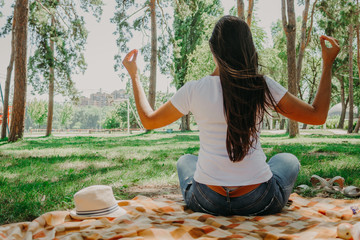 The concept of harmony with nature, meditation in the fresh air. A girl with long black hair in a white t-shirt and jeans is sitting on the grass in a Park among pine trees, in a yoga pose, rear view.