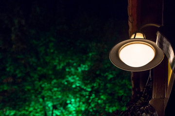 an iron lamp with a glass frosted shade illuminates the street with warm light on the facade of the building against the background of green leaves of trees in the night.