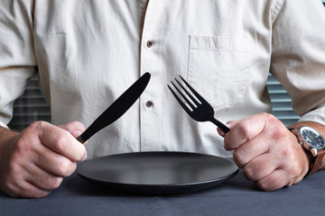 A man in a beige shirt at a table with a black empty plate. Black fork and knife.