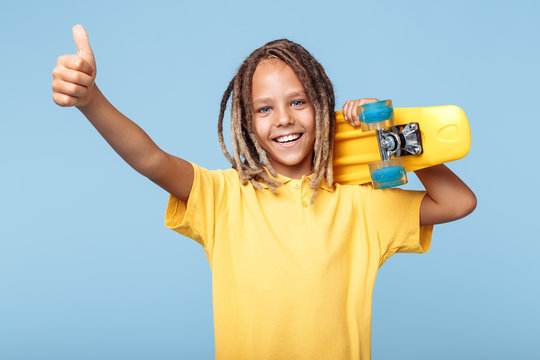 Positive little boy with african dreads holding skateboard on shoulders and showing thumb up over blue background.