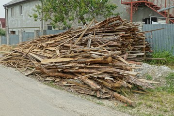 a pile of firewood from long wooden planks and logs on the street near the fence near the asphalt gray road
