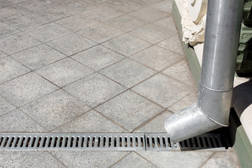 an engineering structure a downspout with a drainage grate against a background of a gray stone granite sidewalk from square tiles with copy space, side view closeup nobody.