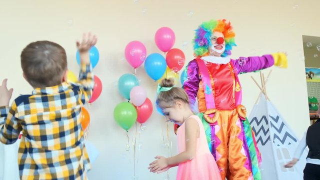 cute little friends and funny female clown in bright suit dance in decorated room with wigwam at birthday party slow motion