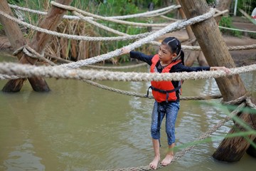 A cute young Asian girl with an orange life vest is going through an obstacle course at a summer camp with tight rope hanging over water.