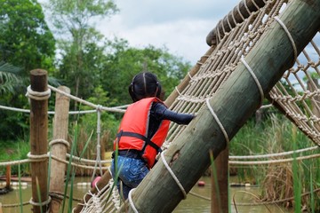 A cute young Asian girl wearing orange life vest is going up a climbing net in an obstacle course at a summer camp.