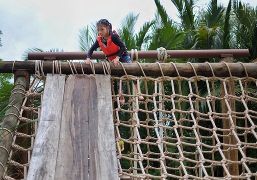 A cute young Asian girl wearing orange life vest is going up a climbing net in an obstacle course at a summer camp.