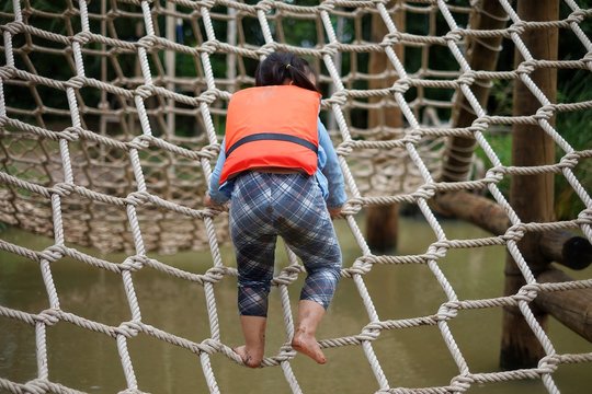 A cute young Asian girl wearing orange life vest is going up a climbing net in an obstacle course at a summer camp.