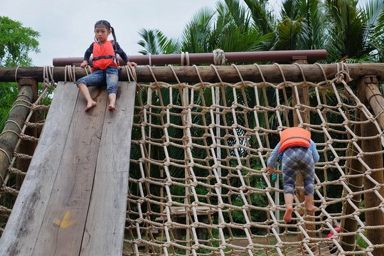 Two cute young Asian girls wearing orange life vest are going up a climbing net in an obstacle course at a summer camp.